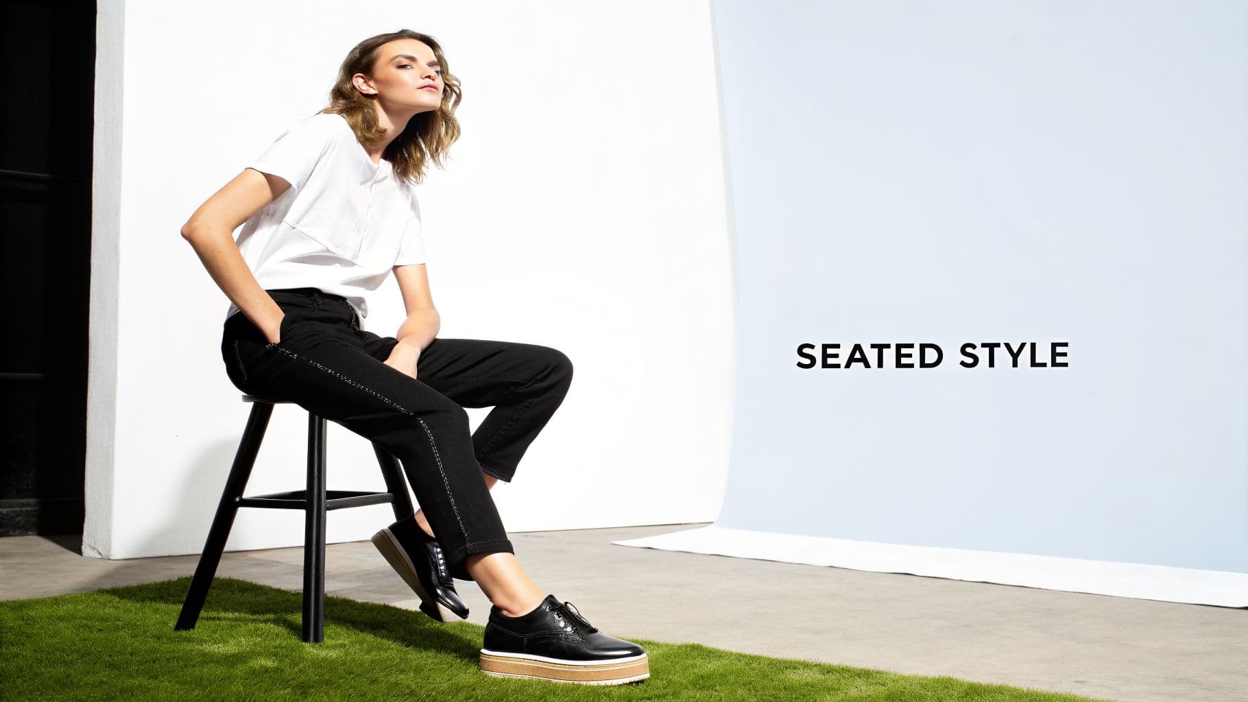 A stylish woman in a white t-shirt and black jeans poses on a stool with platform shoes.