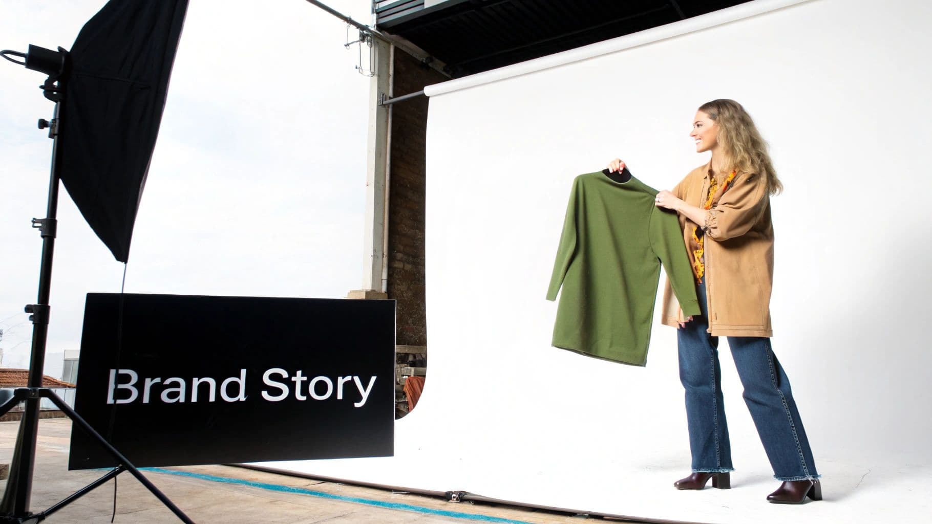 A smiling woman holds a green sweater during a fashion photoshoot in a studio with a 'Brand Story' sign.